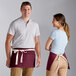 A man and woman wearing Choice burgundy waist aprons with natural webbing standing in a professional kitchen.