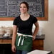 A woman wearing a Choice hunter green waist apron with natural webbing standing in front of a chalkboard.
