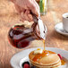 A hand using a Vollrath syrup dispenser to pour syrup on pancakes on a table.