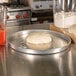 A dough on an American Metalcraft aluminum pizza pan next to a container of flour and a blender.