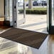 A large brown Lavex Needle Rib entrance mat in front of a building's glass doors.