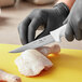 A person in black gloves using a Choice 5" wide stiff boning knife to cut up a chicken on a counter.