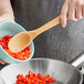 A person using an OXO small wooden spoon to stir chopped red peppers in a bowl.