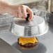 A hand holding an American Metalcraft aluminum basting cover over a hamburger on a grill.