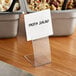 A clear plastic Choice deli tag holder with a white label on a table next to food.