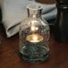 A Sterno clear glass lantern candle holder with a lit candle inside sitting on a table.