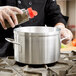 A chef pouring seasoning into a Vollrath classic aluminum stock pot on a stove.