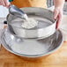 A person pouring flour into a Choice stainless steel sieve over a bowl.