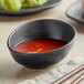A dark gray Riverstone melamine ramekin filled with red sauce on a table with chopsticks.