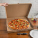 A hand holding a 20" white corrugated pizza circle over a table with a pizza in a box.