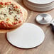 A white pizza with mushrooms on a white 9" corrugated pizza circle next to a pizza cutter.