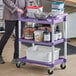 A woman standing next to a purple and white Choice utility cart with food containers on it.