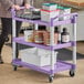 A woman pushing a purple Choice utility cart with three shelves and a variety of containers on it.