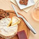 A Boska stainless steel soft cheese knife cutting into a round white cheese on a table with other cheese and bread.