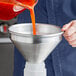 A person pouring orange liquid into an American Metalcraft aluminum funnel.