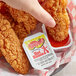 A hand holding a Texas Pete Original Hot Sauce dip cup next to fried chicken strips.