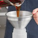 A person pouring brown liquid into an American Metalcraft aluminum funnel.