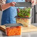 A woman using a Vigor 1/3 size clear plastic food pan with a secure sealing cover to hold salad and shredded carrots.