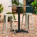 A Lancaster Table & Seating Excalibur outdoor table base with a bar height column and white stools on a brick patio.