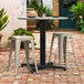 A Lancaster Table & Seating Excalibur outdoor table with FLAT Tech table levelers on a brick patio with two white stools.