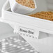 A white Baker's Mark ingredient bin with brown rice in it and a white spoon.