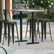 A Lancaster Table & Seating rectangular bar table with smooth finish and two end plates on a patio with black chairs and stools.