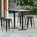 A Lancaster Table & Seating rectangular counter height table with smooth finish and two end outdoor base plates on a patio with stools.