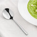 An Acopa stainless steel bouillon spoon next to a bowl of green soup on a white surface.