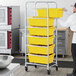 A man in a kitchen wearing an apron and holding a Regency mobile aluminum lug rack full of yellow tote boxes.