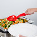 A close-up of a hand holding a red Cambro perforated salad bar spoon over a tray of vegetables.