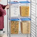 A woman standing next to a Vigor polycarbonate food storage container full of pasta on a metal shelving unit.