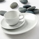 A Tuxton bright white round china cup on a saucer on a white surface with rocks around it.