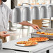 A man in a white shirt and black hat uses a ServIt suspension bar to serve himself food from a counter with pizza.