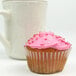 A cupcake with pink frosting and sprinkles in a white fluted baking cup next to a white mug.