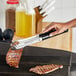 A hand using Choice black and silver scalloped tongs to pick up cooked meat over a grill.