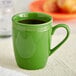 A close-up of a green Acopa Capri stoneware mug with a handle on a white table