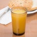 A light amber Cambro Del Mar plastic tumbler filled with orange juice on a table next to a bagel.