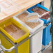 A person using a white scoop to pour brown grains into a blue, white, and yellow Baker's Mark triple ingredient bin.
