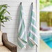 Two Monarch Brands Aston & Arden green and blue striped pool towels hanging on a wall.