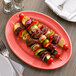 A Tuxton Cinnebar oval china platter with meat and vegetables on skewers on a table.