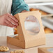 A person opening a brown Kraft paper bakery box with a window to reveal cookies inside.
