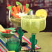 Two green Libbey cactus margarita glasses filled with margaritas on a table with a bowl of guacamole and vegetables.