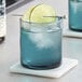 A blue Acopa Pangea rocks glass with a drink and a slice of lime on the table.