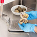 A person in blue gloves using a Vollrath Oyster Shucker to open an oyster on a cutting board.