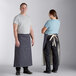 A man and woman standing next to each other wearing Acopa Kennett blue denim bistro aprons with natural webbing.