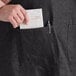 A man wearing a black denim Acopa Kennett restaurant apron with 3 pockets puts a receipt in a pocket.