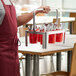 A person in a red apron holding a Choice stainless steel flatware carrier with red and silver utensil holders inside.