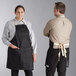 A man and woman wearing Acopa black denim bib aprons with natural webbing standing in a professional kitchen.