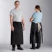 A man and woman standing next to each other wearing Acopa black bistro aprons.