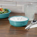 A Fiesta turquoise china bowl filled with salad with white sauce on a table with a glass of liquid.